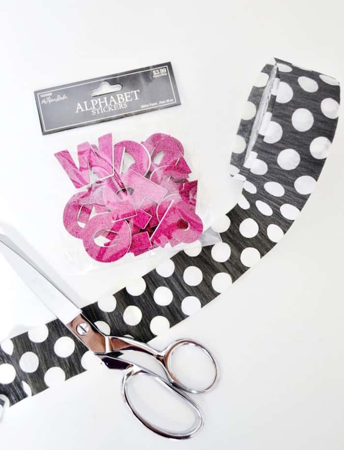 Black and white polka dot crepe paper roll, scissors, and dark pink glitter letters in a bag