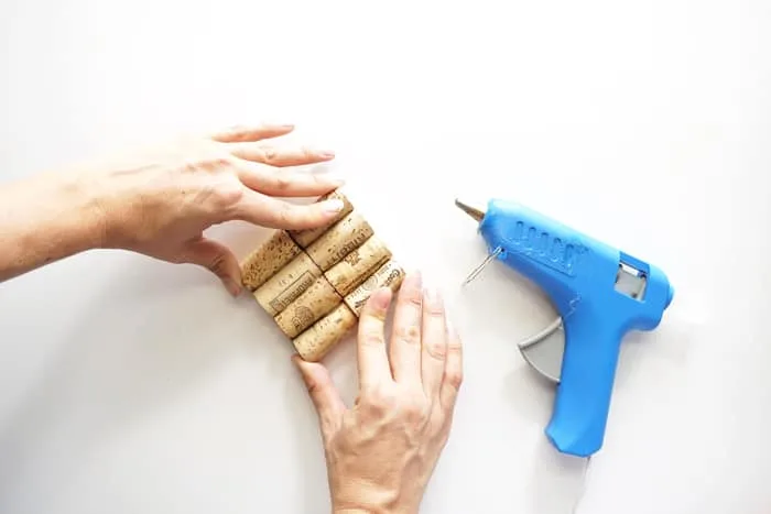 Two hands holding a small wine cork board with a blue hot glue gun