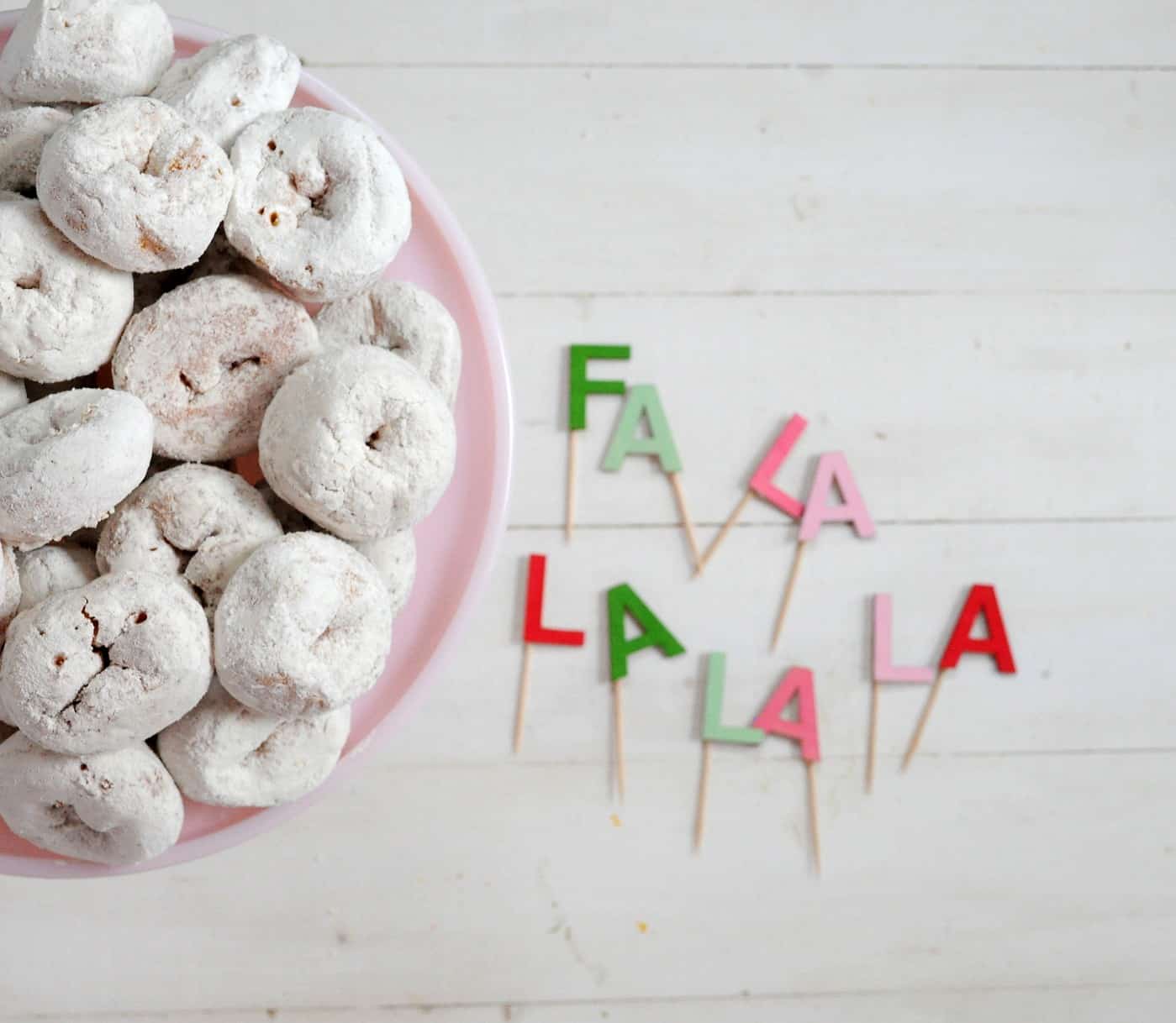 Christmas cake topper that says Fa la la la la next to a plate of white powdered donuts