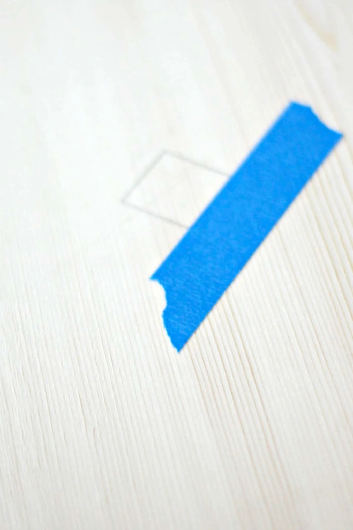 Piece of painter's tape next to a square drawn in pencil on a wood board
