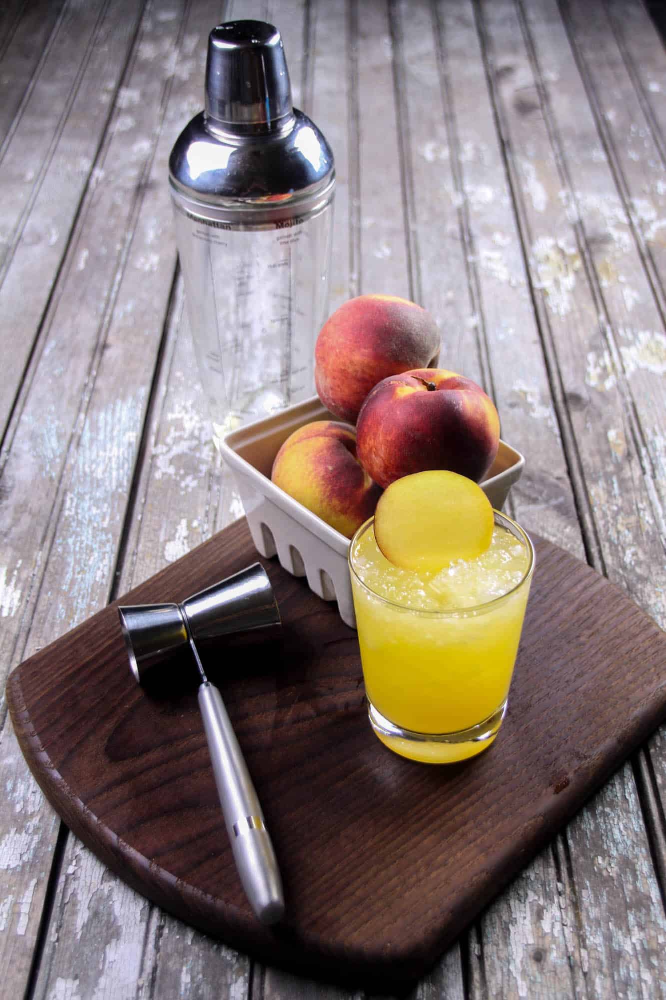 Cocktail shaker along with a basket of peaches and a peach vodka cocktail