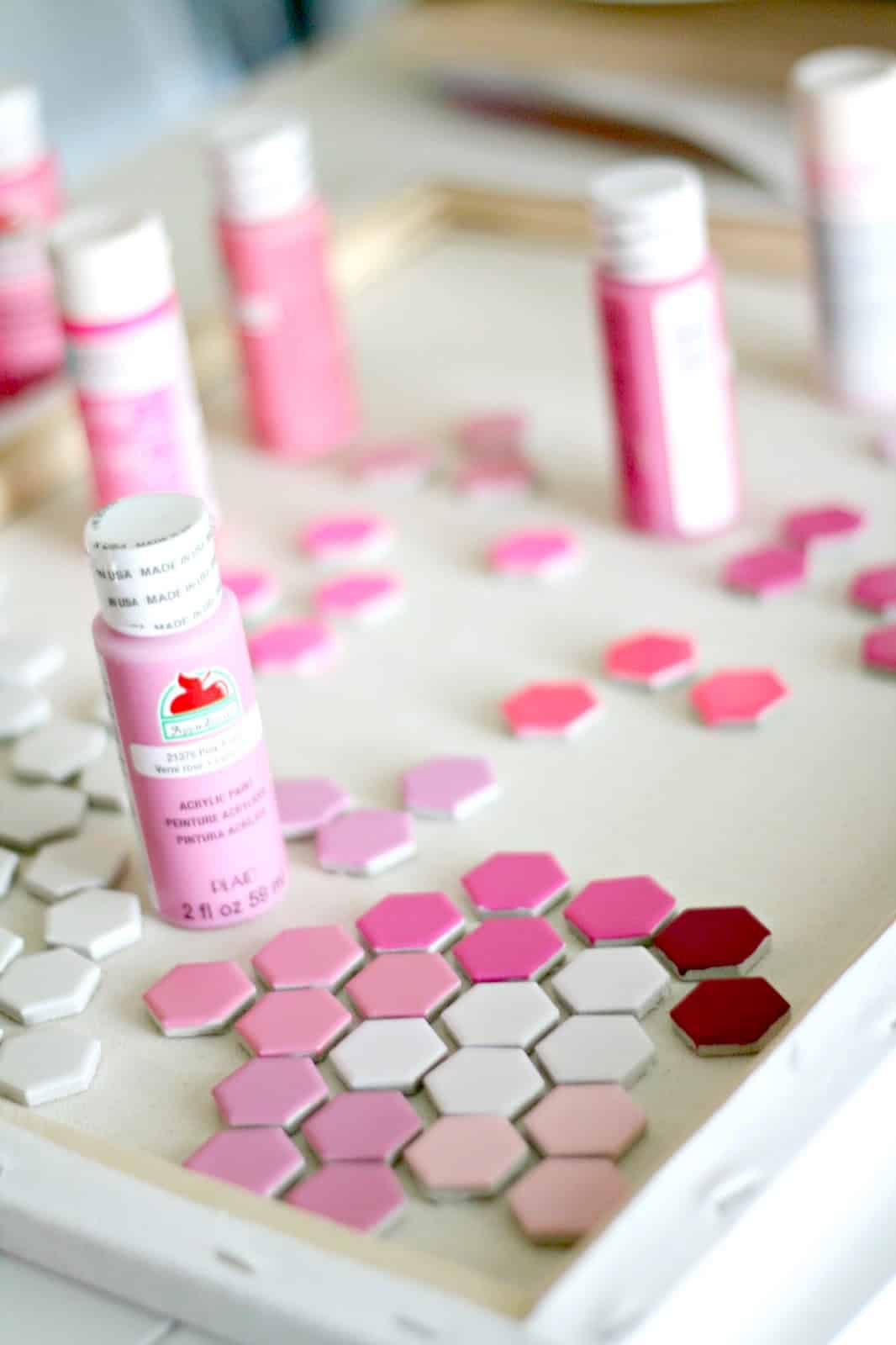 Hexagon tiles painted with various shades of pink and red paint with paint bottles in the background