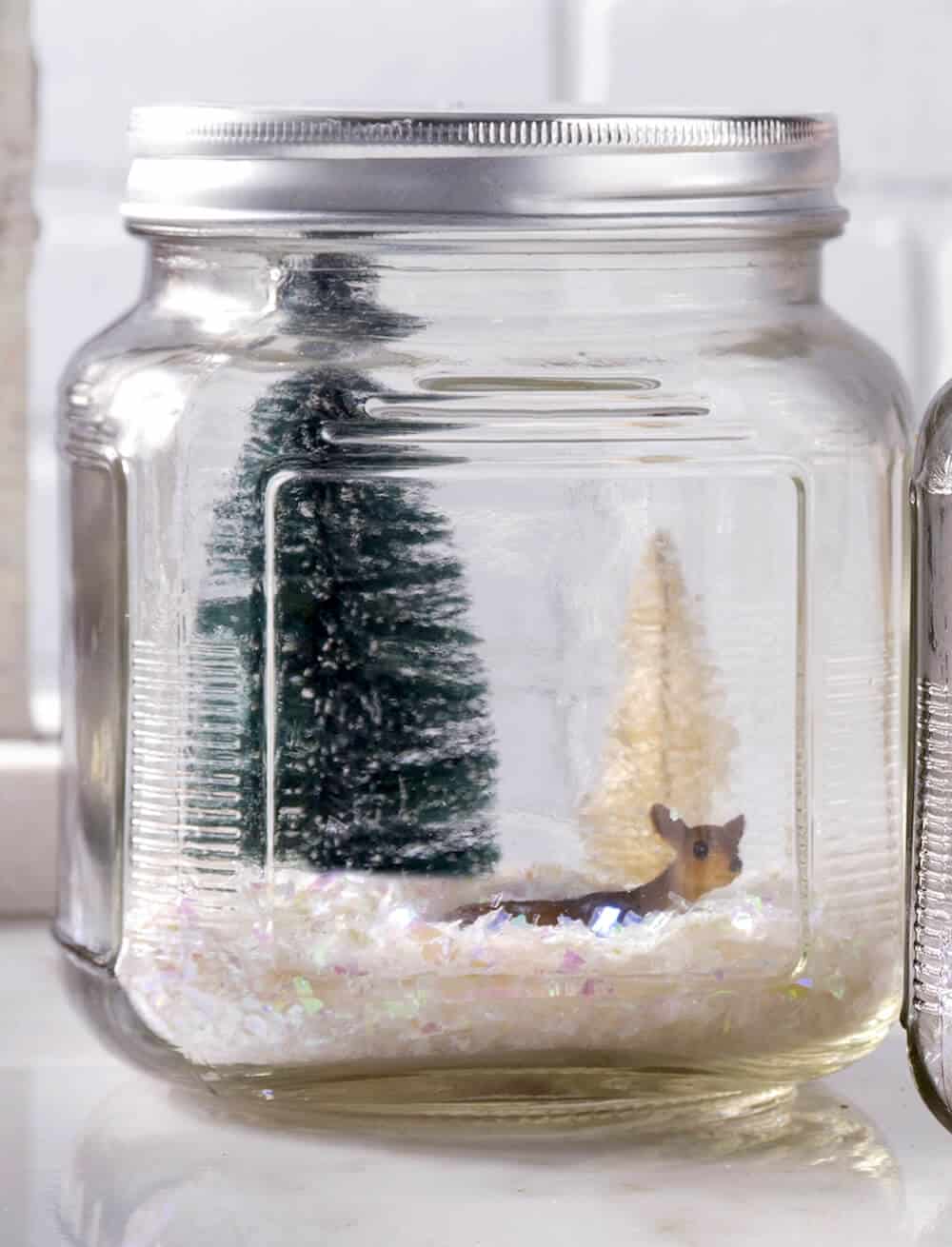 Winter wonderland glass jar filled with artificial snow, bottle brush trees, and a plastic deer