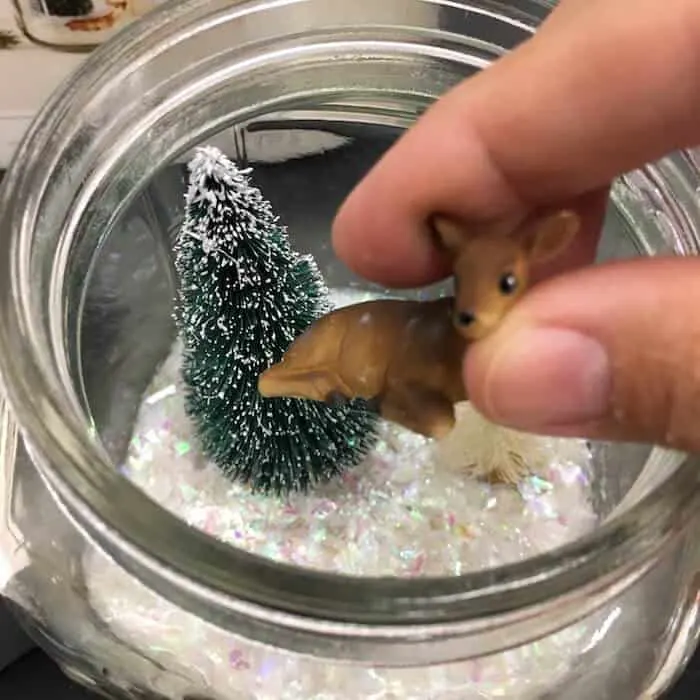 Placing plastic animals into a glass jar filled with bottle brush trees and artificial snow