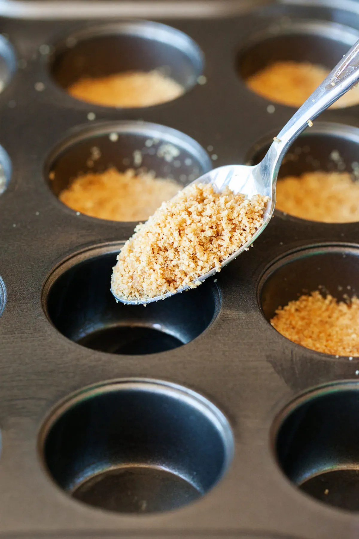 Pouring crushed up graham cracker crumbs into a muffin tin with a spoon