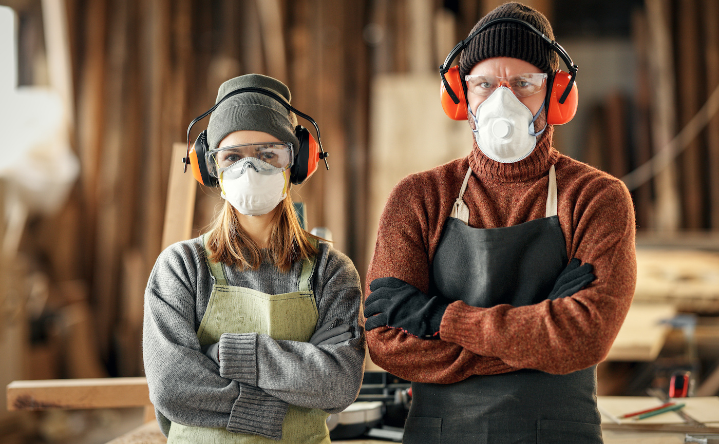 Couple of professional carpenters in respirators and protective goggles and headphones looking at camera while standing together in workshop