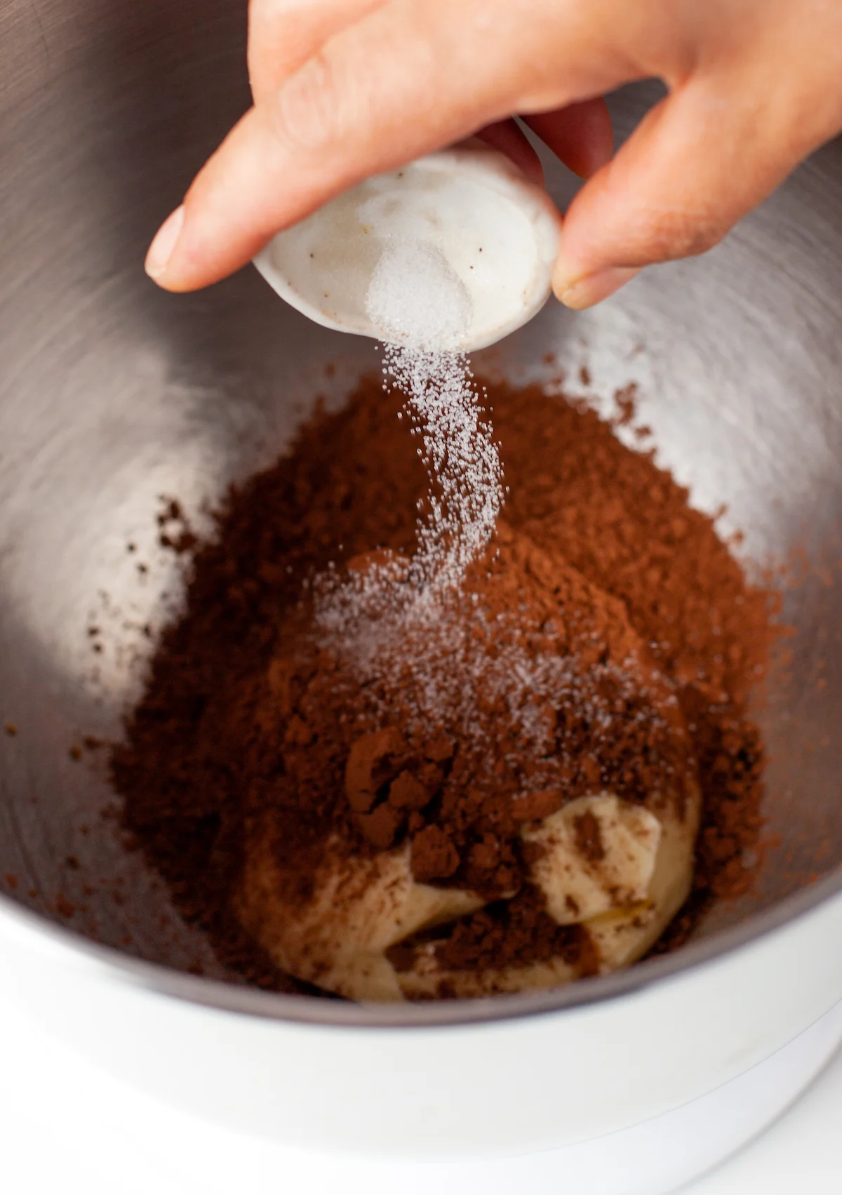 Salt being poured into a metal mixing bowl with cocoa powder and butter